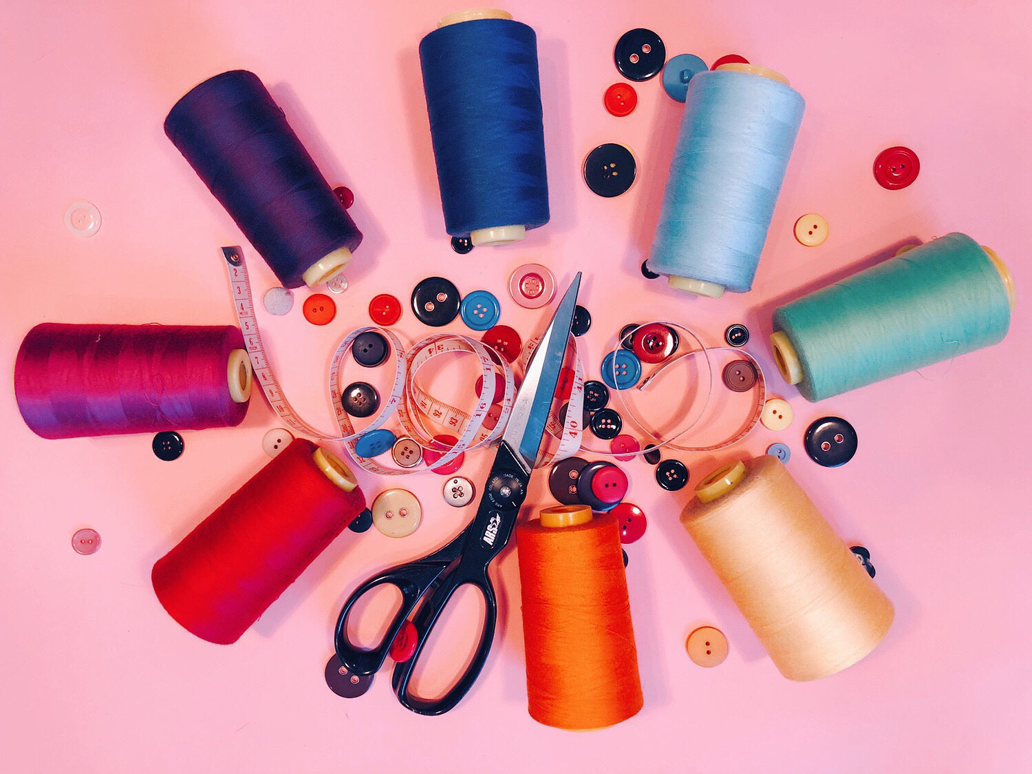 colorful rolls of thread buttons and scissors on pink background