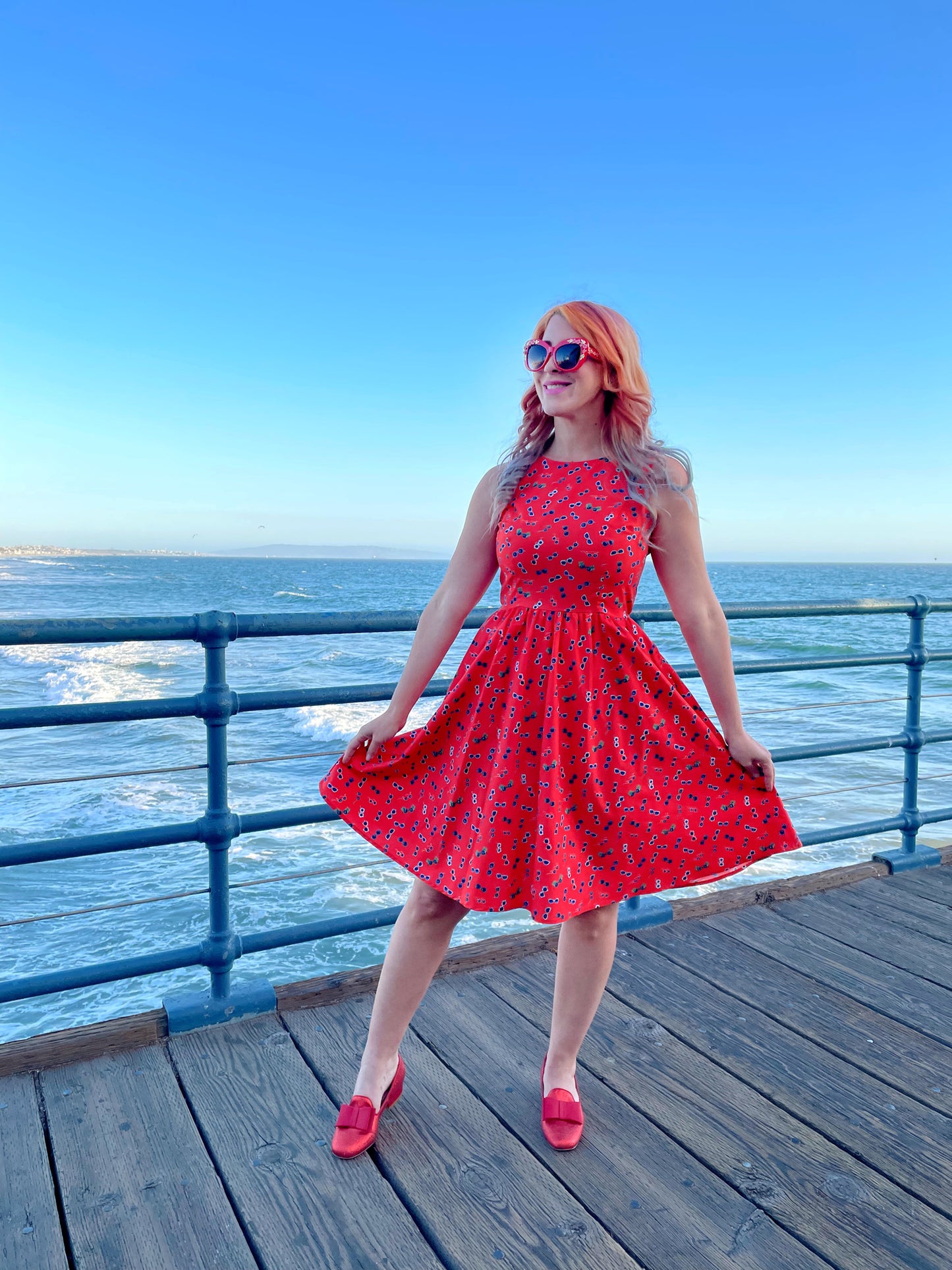 a model standing on the boardwalk in front of the ocean wearing sunglasses vintage dress and red sunglasses