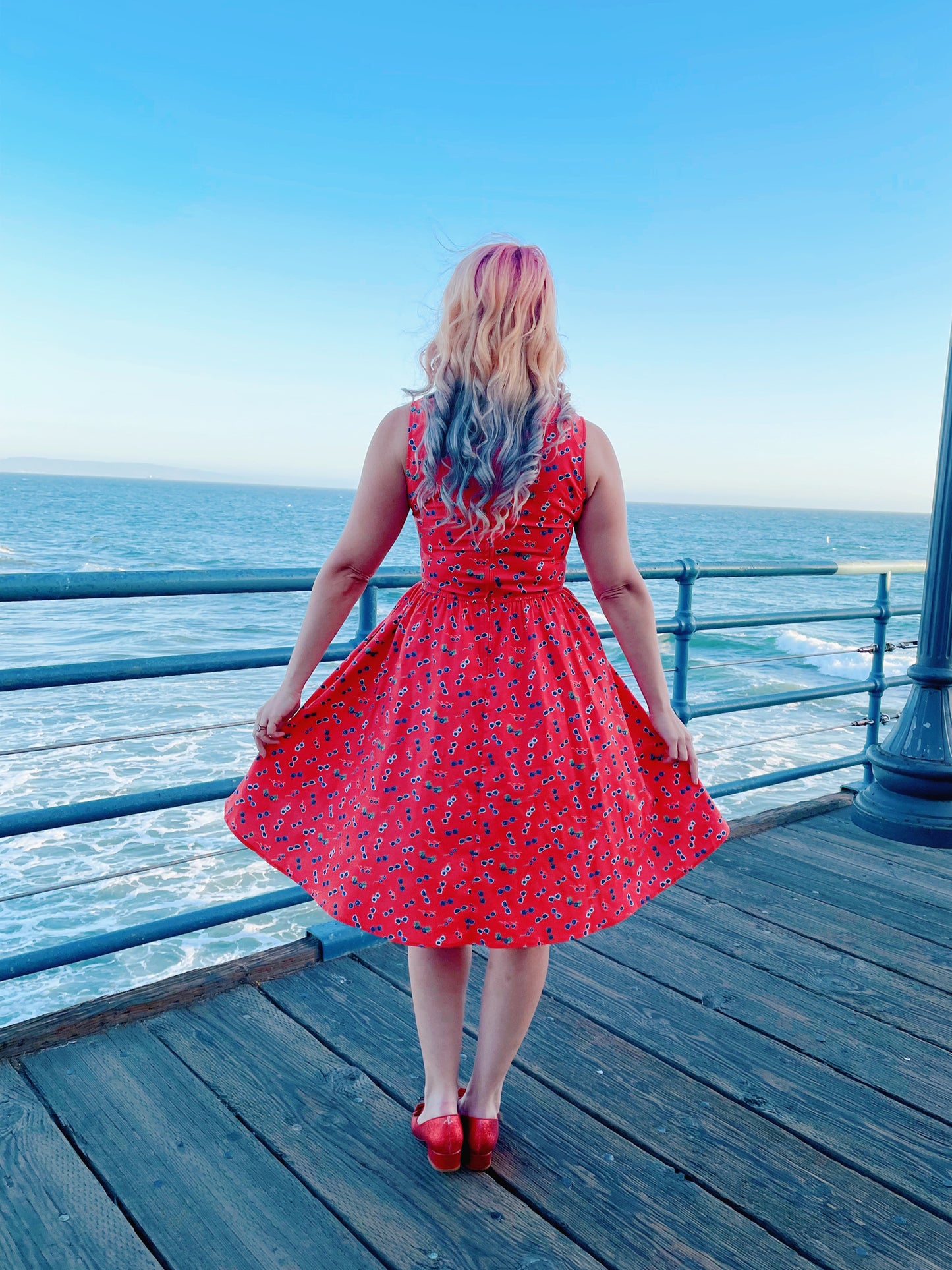 a back view of model standing on the boardwalk in front of the ocean wearing sunglasses vintage dress and red sunglasses