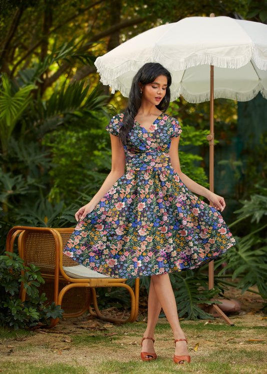 Woman in a floral dress standing outdoors with a white umbrella and wicker chair in the background.