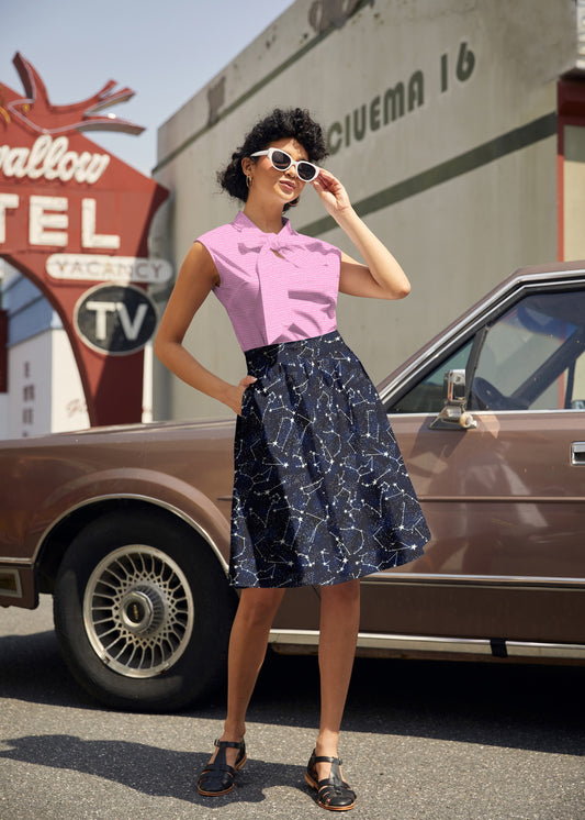 Woman in a pink top and navy skirt standing next to a vintage car with a motel sign in the background.