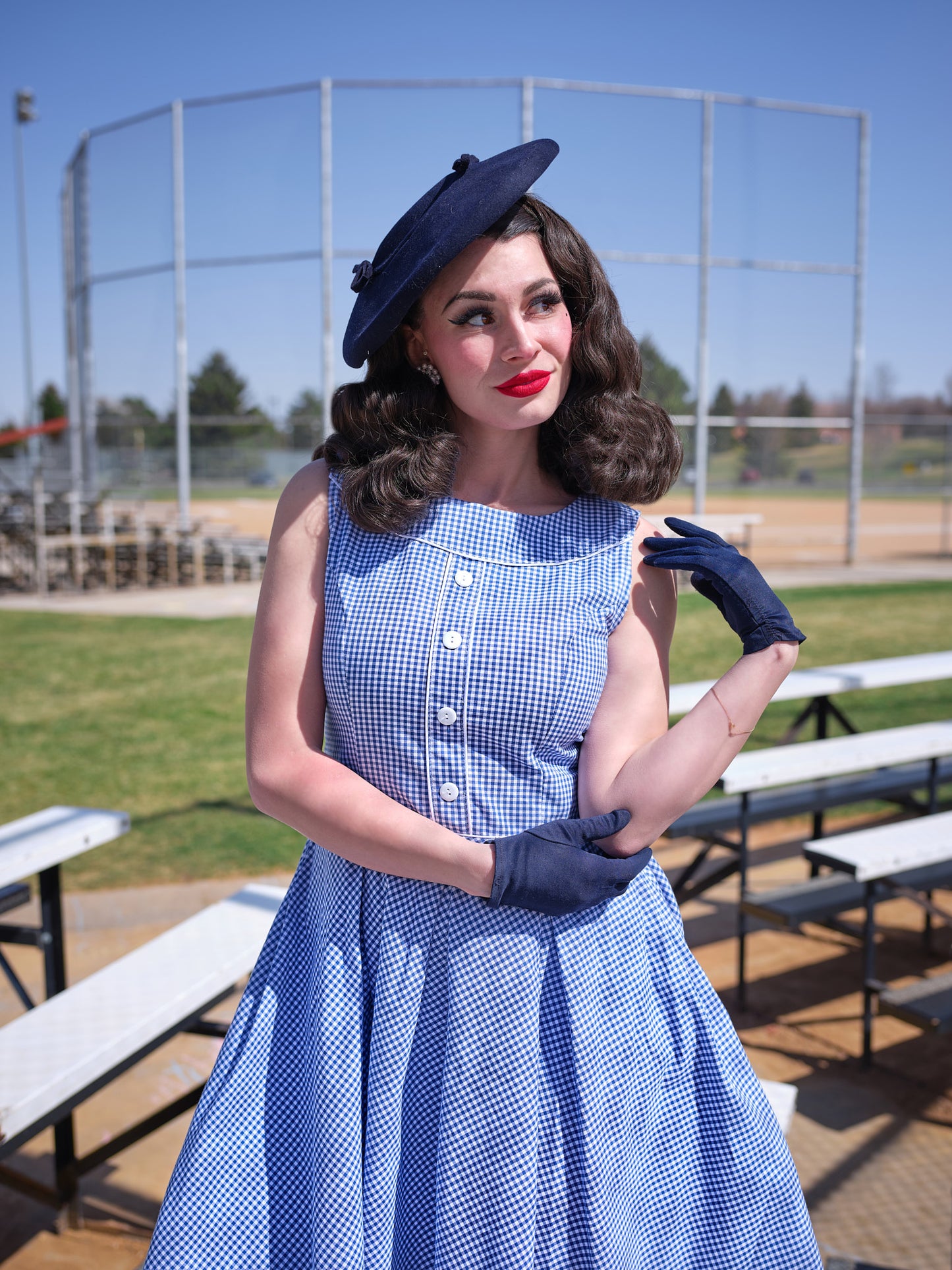 a close up of a model wearing elizabeth dress in gingham and vintage hat and gloves standibg in a baseball field