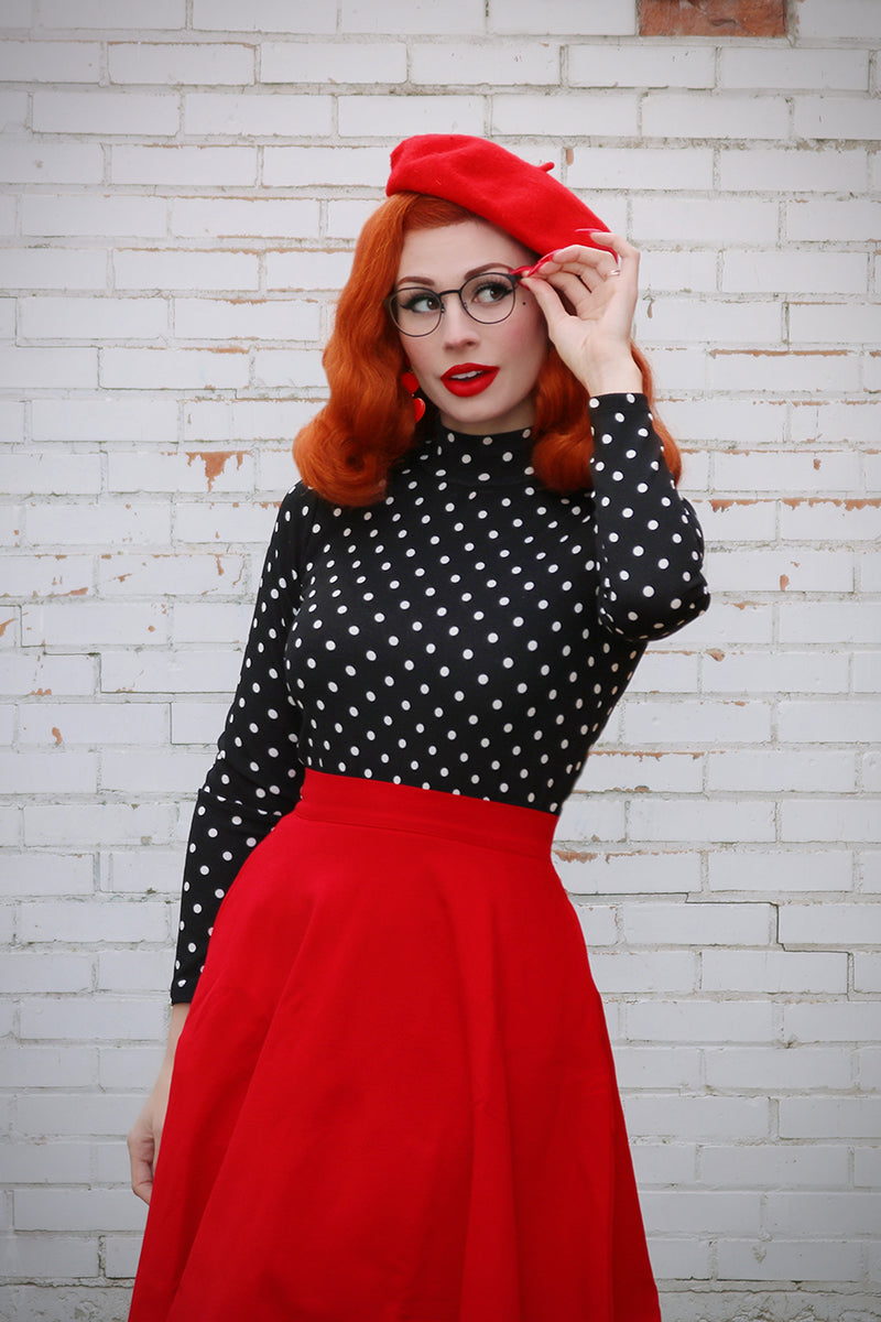 Model posing while wearing a long red skirt, black top with white polka dots and red beret.
