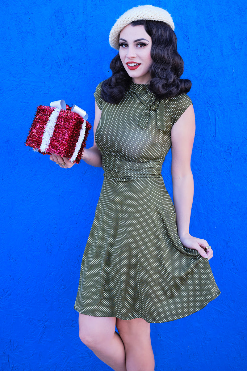 a model standing in front of blue wall hodling a gift wearing olive dot bombshell dress and white beret styled in vintage fashion 