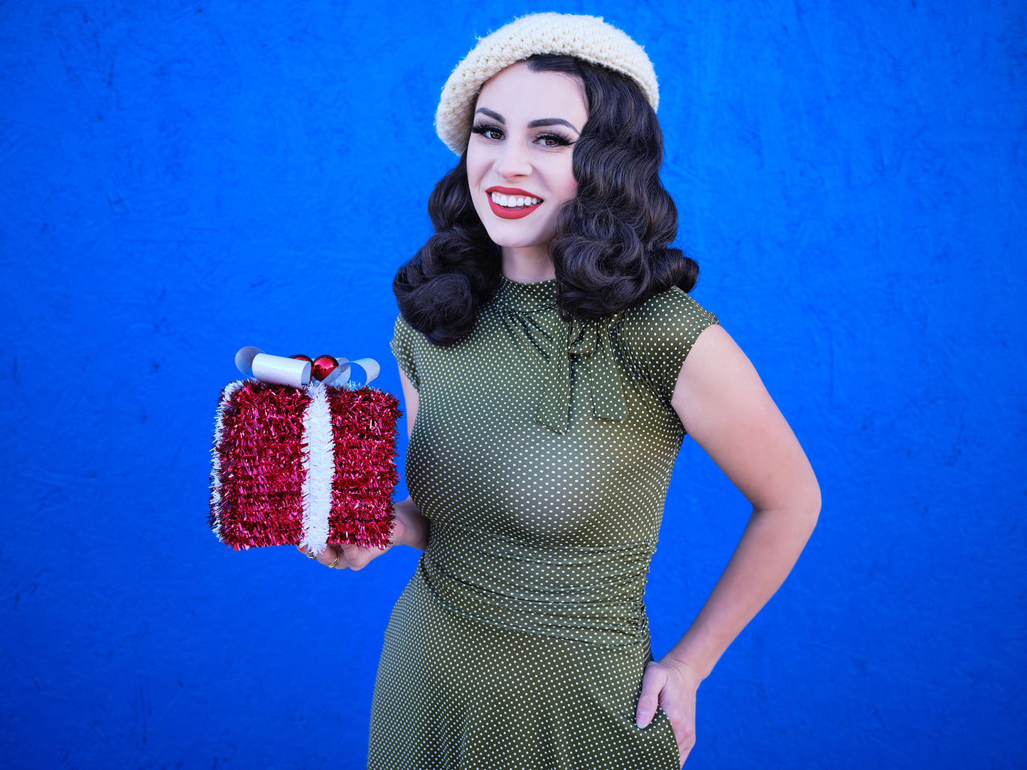 a close up of a model standing in front of blue wall hodling a gift wearing olive dot bombshell dress