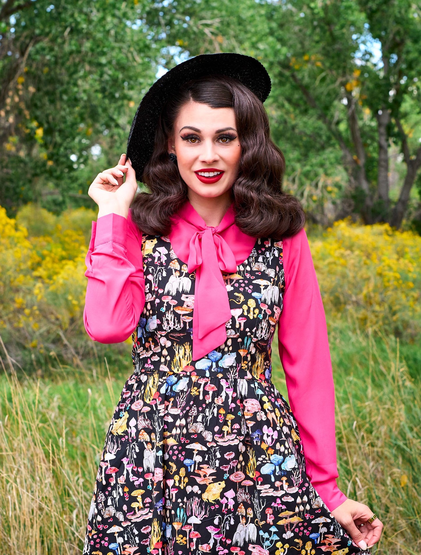 a close up image of a model standing in front of trees wearing the helen retro blouse in pink underneath a dress