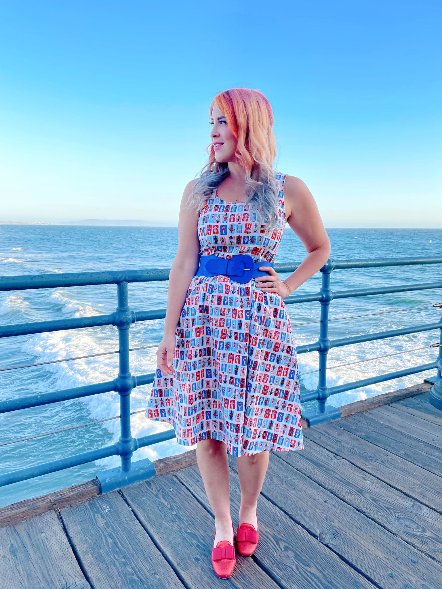 a model standing on a boardwalk with the ocean in the backgroun wearing the sunny days fit and flare dress