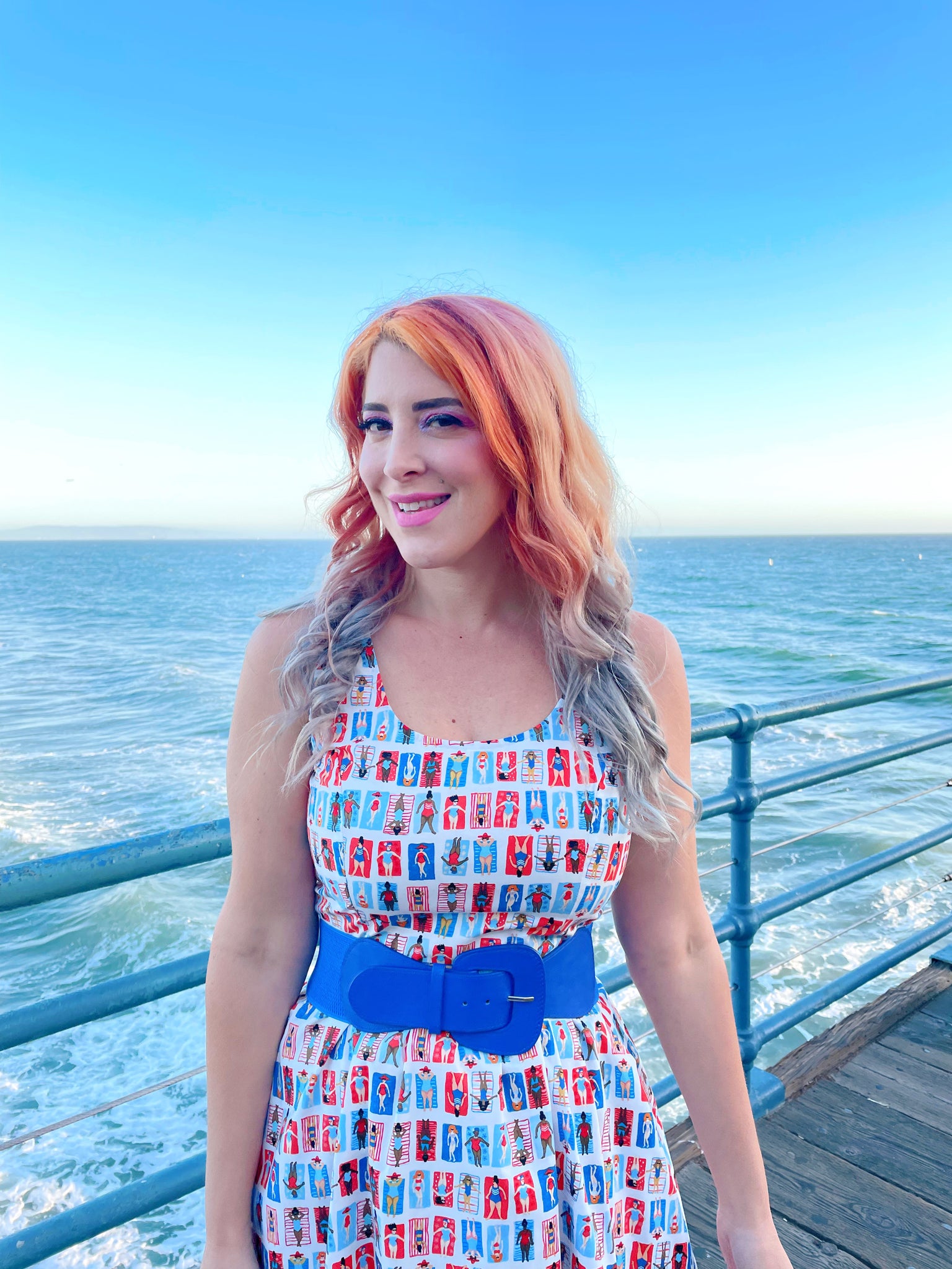 a close up of model standing on a boardwalk with the ocean in the backgroun wearing the sunny days fit and flare dress
