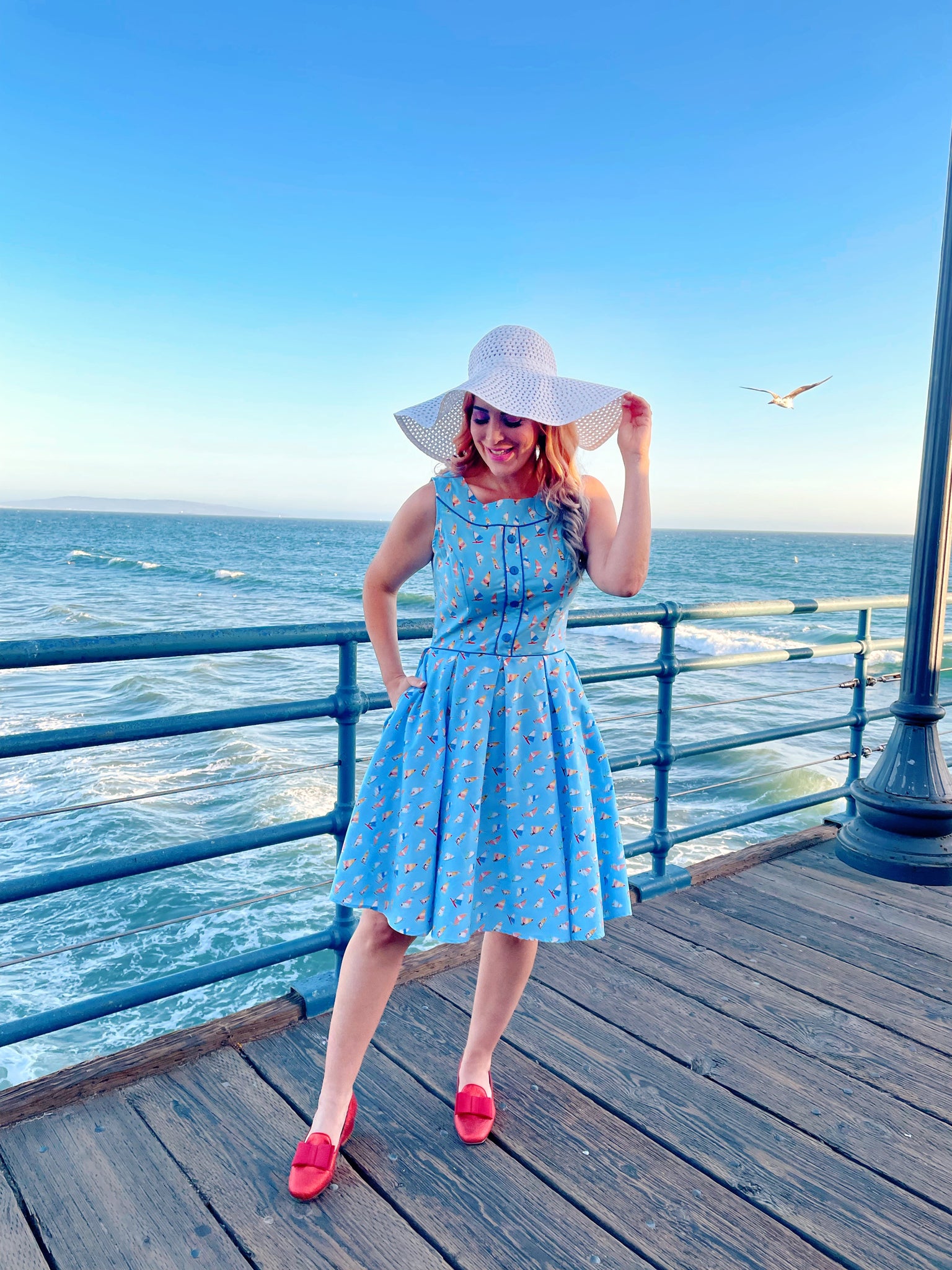 a model standing on the boardwalk in front of the ocean wearing elizabeth dress in windsurfer and white sun hat