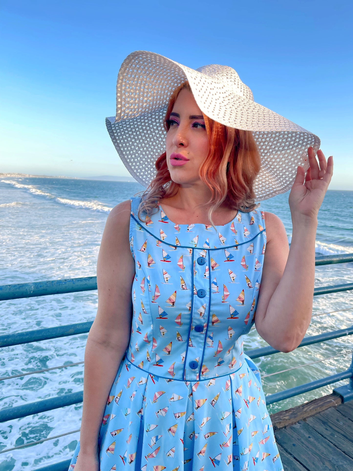 a close up of model standing on the boardwalk in front of the ocean wearing elizabeth dress in windsurfer and white sun hat