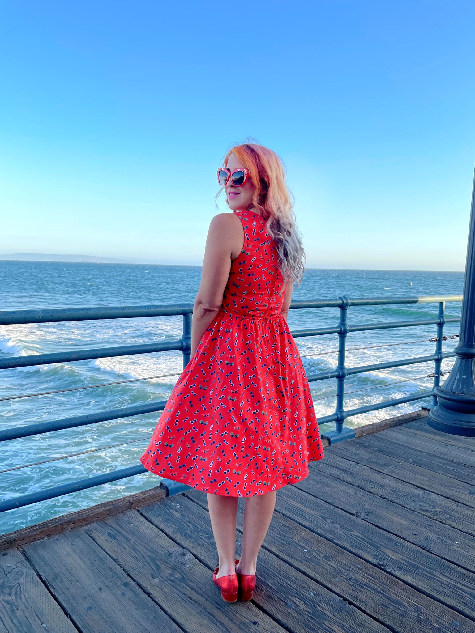 a back view of model standing on the boardwalk in front of the ocean wearing sunglasses vintage dress and red sunglasses