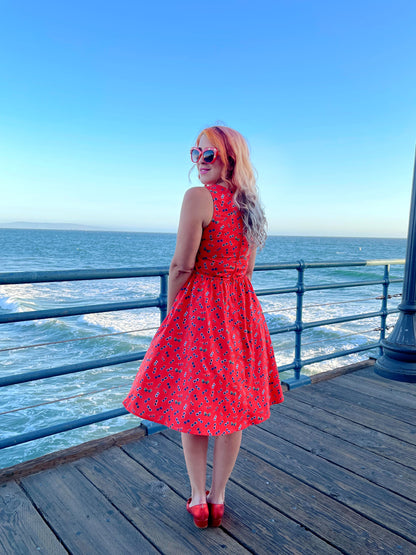 a back view of model standing on the boardwalk in front of the ocean wearing sunglasses vintage dress and red sunglasses