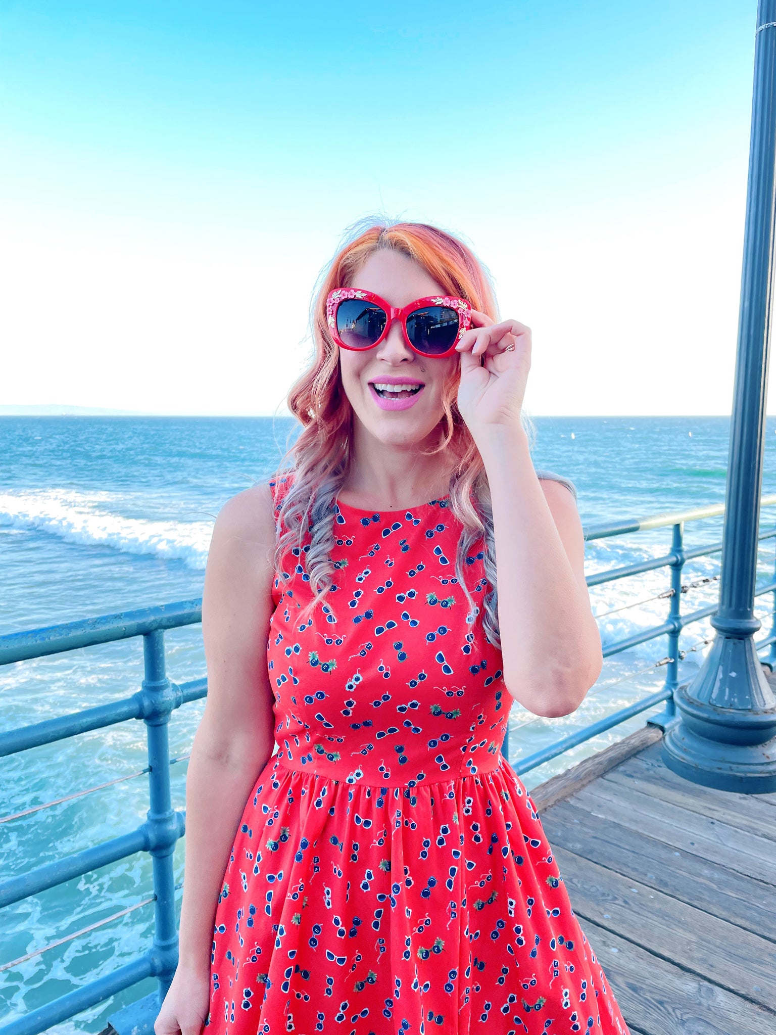 a close up of model standing on the boardwalk in front of the ocean wearing sunglasses vintage dress and red sunglasses
