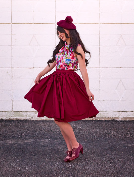 a full size image of a model standing in front of a white wall wearing the peggy swing skirt in burgundy 