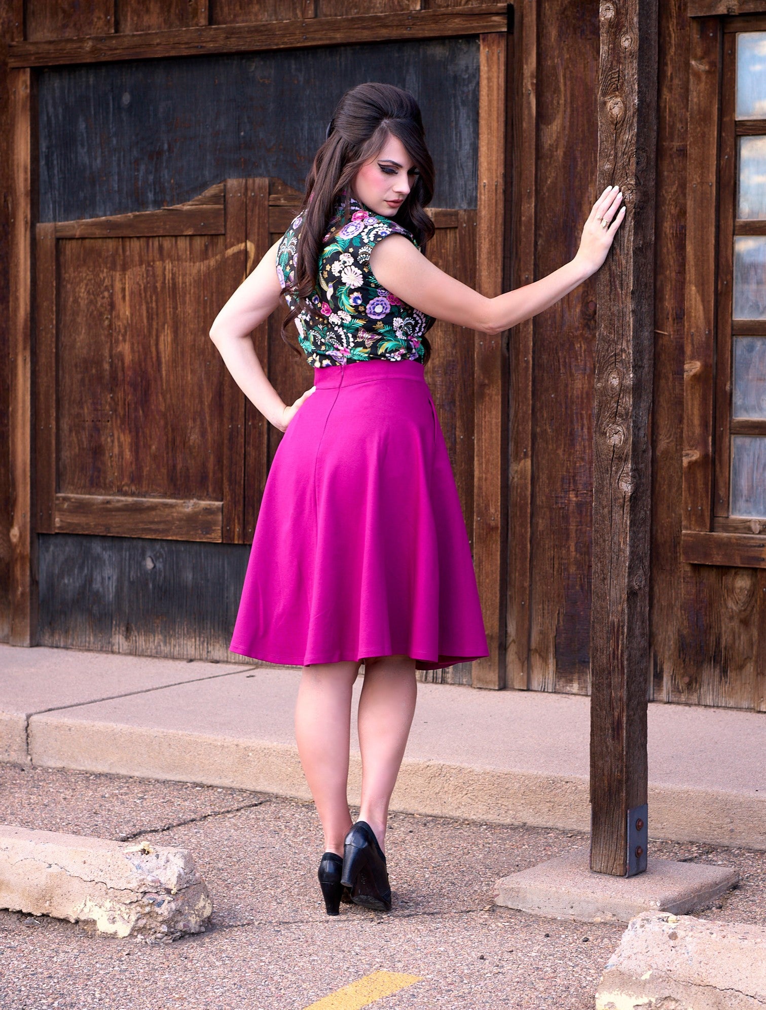 a full size image of the back of a model standing in front of a wooden wall holding a post and wearing the charlotte skirt in magenta