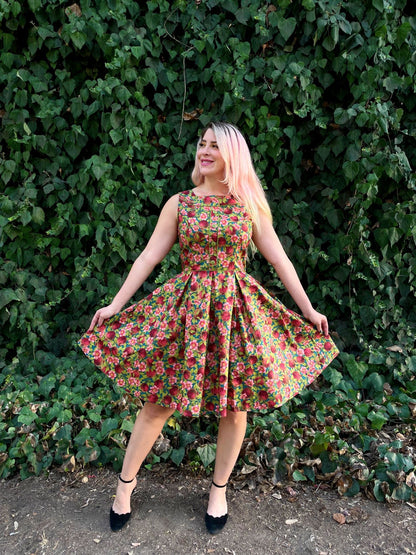 a full length image of a model standing in front of leafy backdrop wearing the Elizabeth dress in pomegranate holding the sides of the skirt