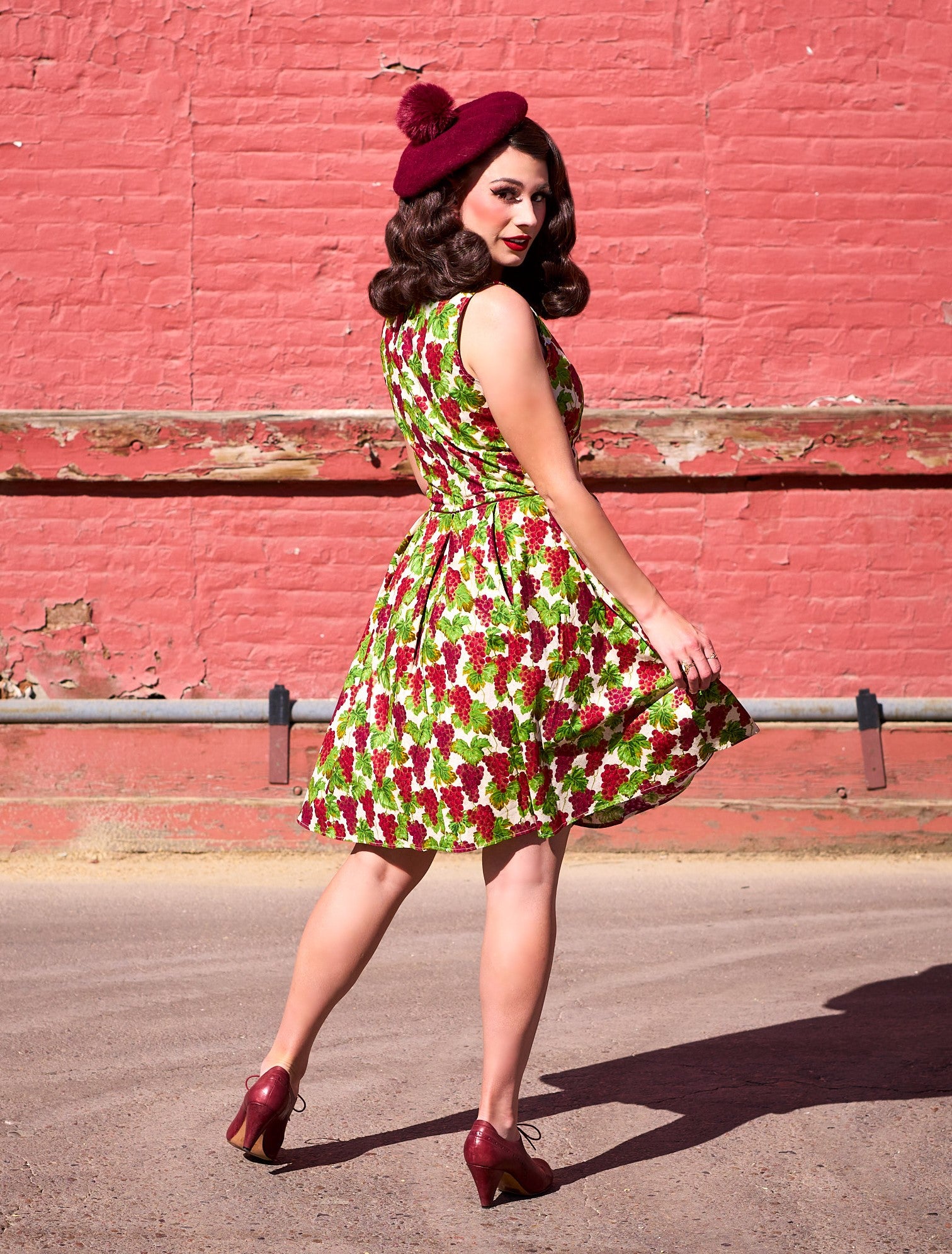 a full size image of the back side a model standing in front a red wall wearing the Elizabeth dress in grapes