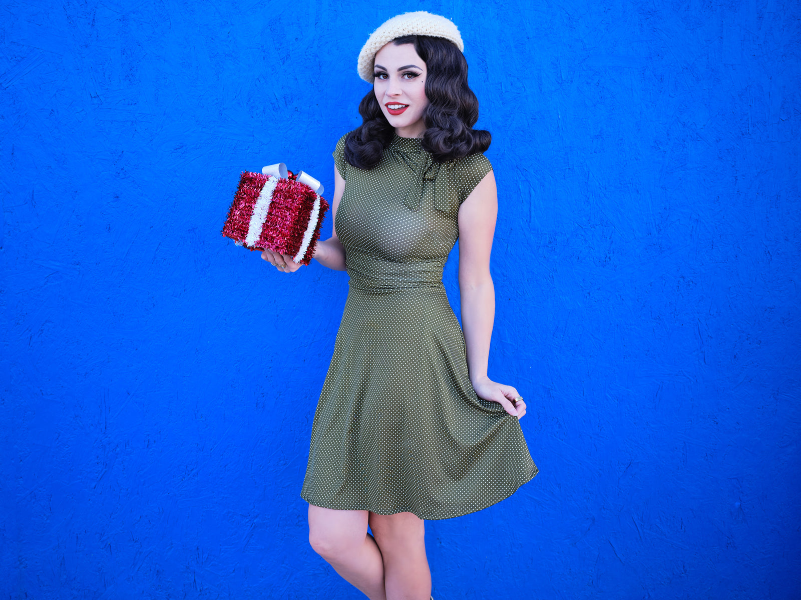 a model standing in front of blue wall hodling a gift wearing olive dot bombshell dress