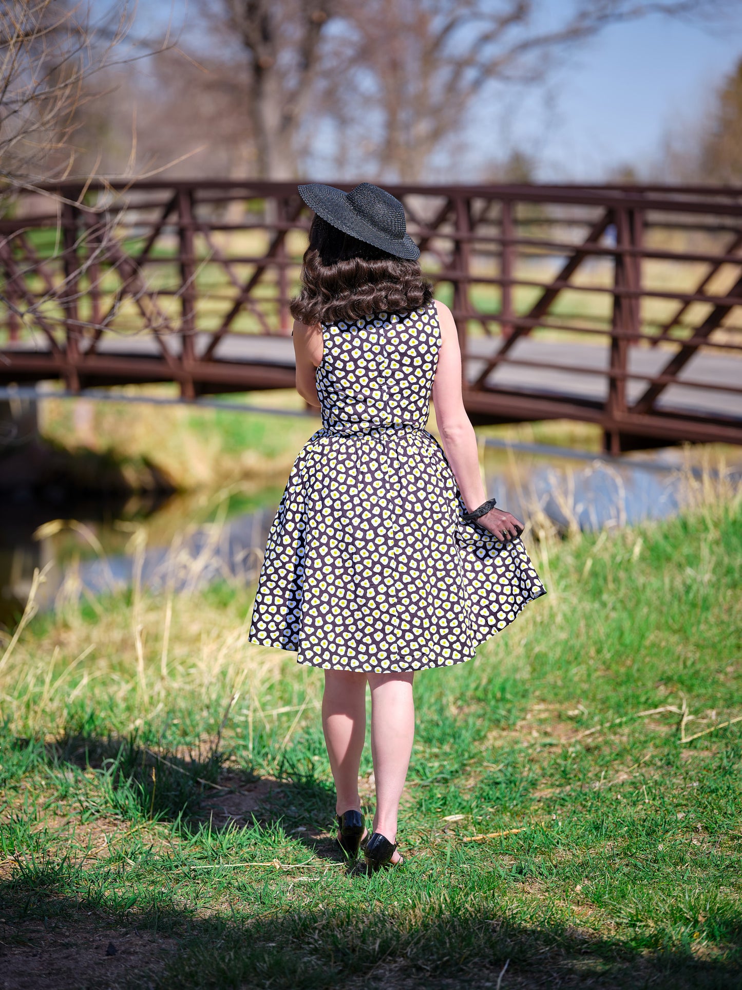 a full size image of a backshot of a model in front of a bridge wearing sunny side up vintage dress and a vintage hat