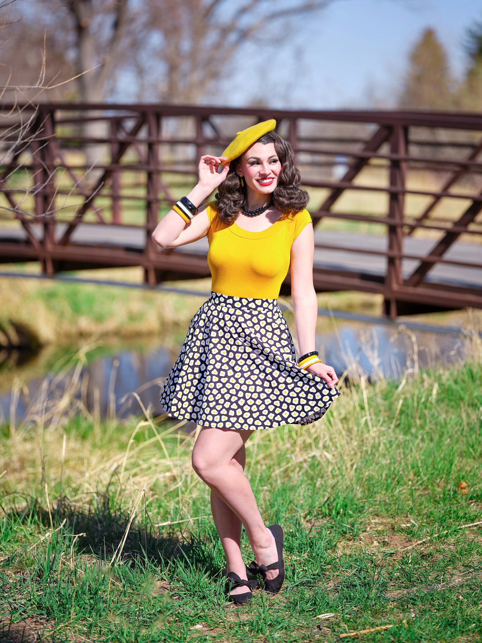 a model standing outdoors in front of a bridge wearing the sunny side up skater skirt with mustard top and beret