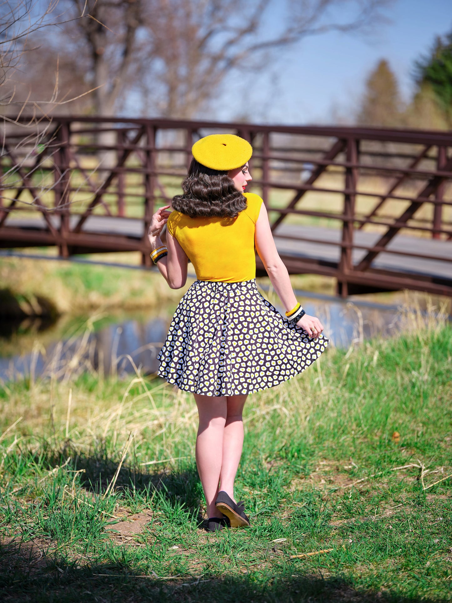 a back view of model standing outdoors in front of a bridge wearing the sunny side up skater skirt with mustard top and beret