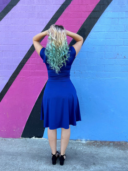 a full length backshot of a model standing in front of a colorful wall wearing bombshell dress in royal blue and holding her hair with both arms