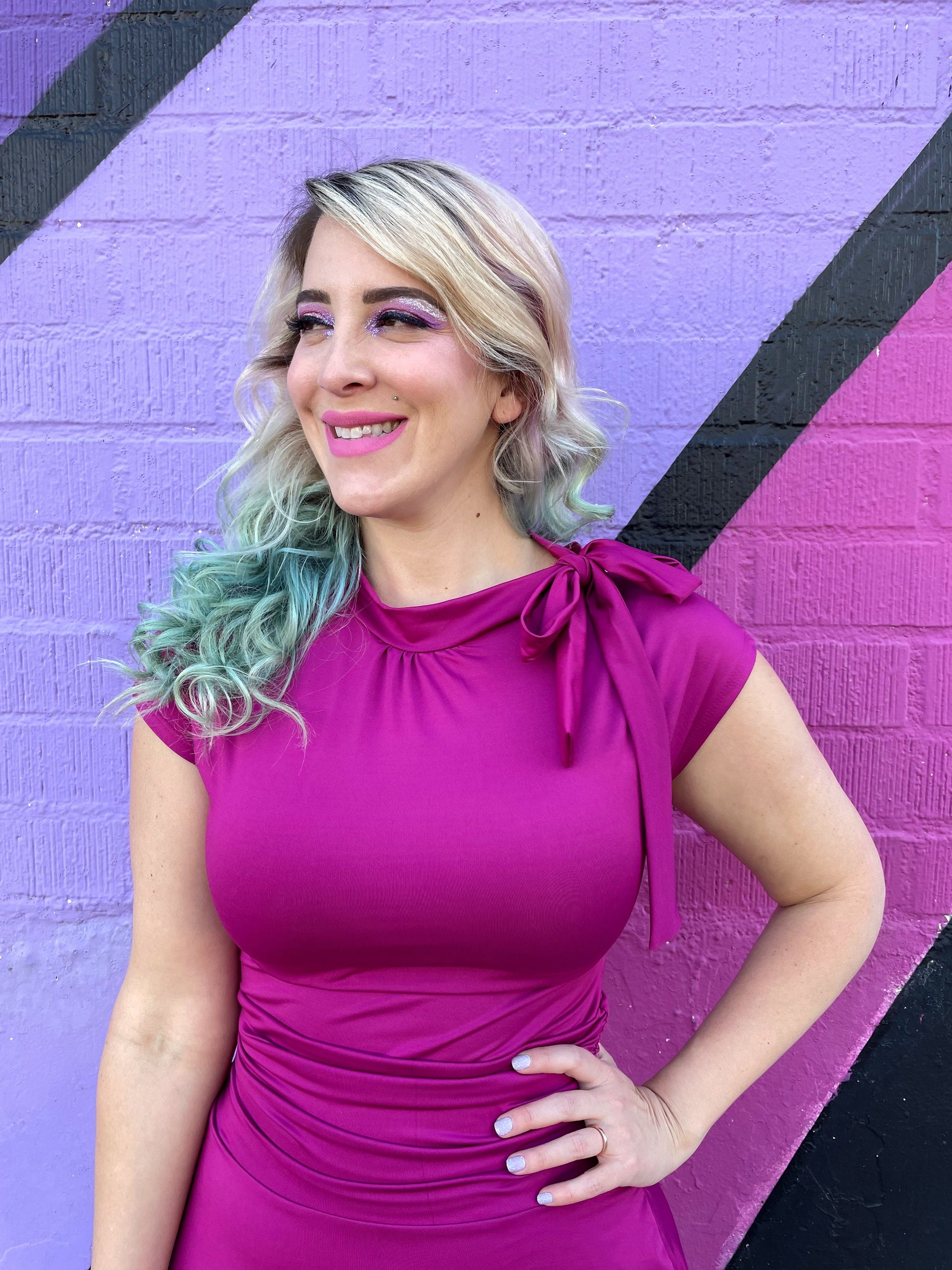 a close up of a model standing in front of a colorful wall with her hand on her hip wearing bombshell dress in magenta