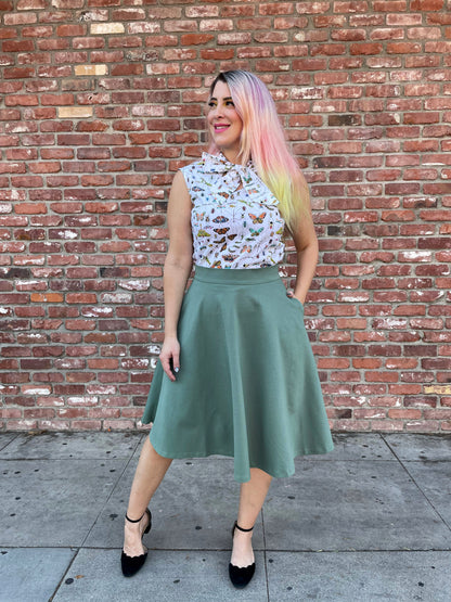 a full size image of a model standing in front of a brick wall wearing the charlotte skirt in sage and her one hand is inside the pocket