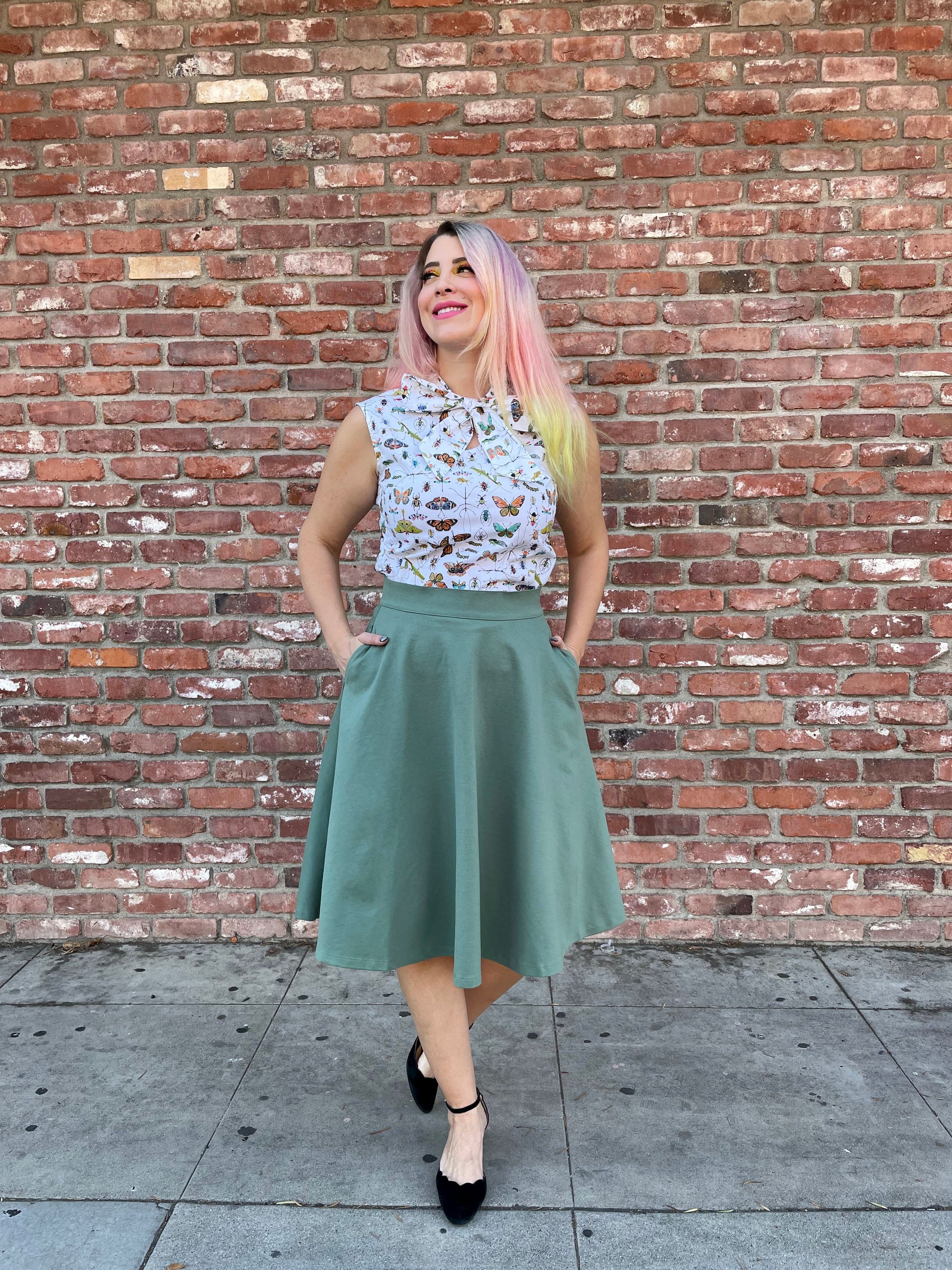 a full size image of a model standing in front of a brick wall wearing the charlotte skirt in sage and her hands are inside the pockets