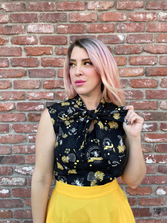 a close up of a model standing in front of a brick wall wearing our bee kind sleeveless bow tie blouse