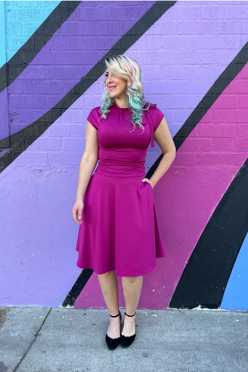a full size of a model standing in front of a colorful wall with her hand in her pocket wearing bombshell dress in magenta